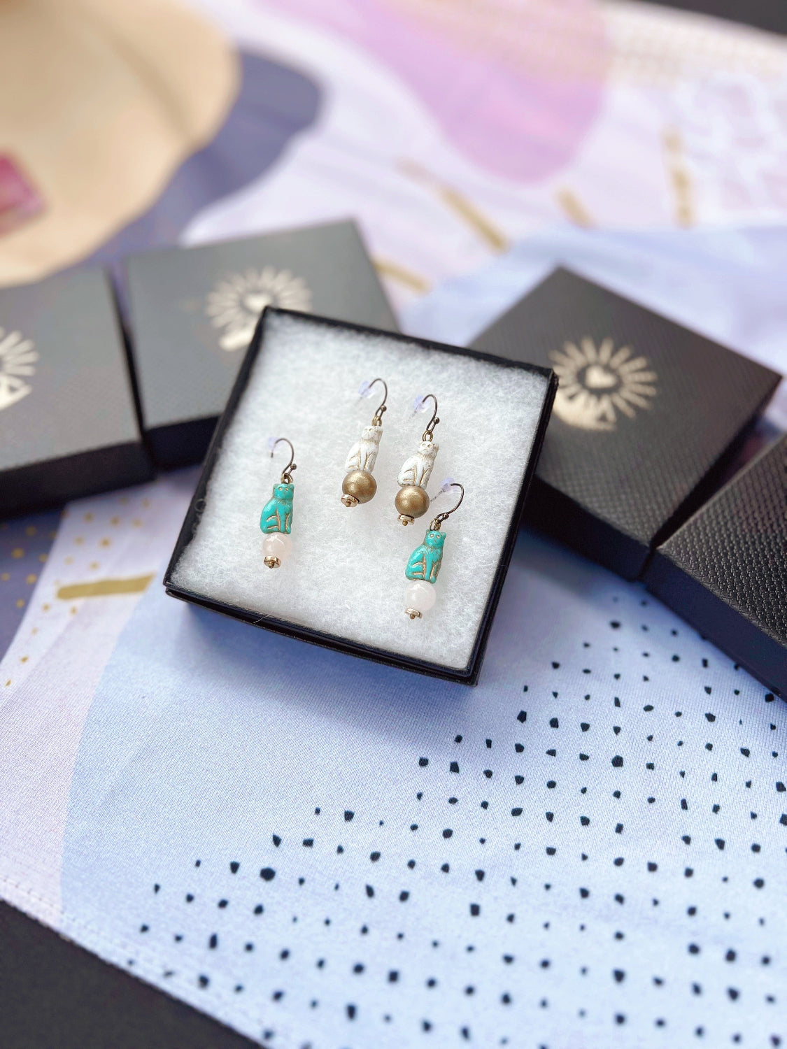 Jewelry boxes with the Oh, Heart! logo spray painted on the top. The boxes are laid across a colorful table runner. One box is open and displaying two pairs of cat dangle earrings.
