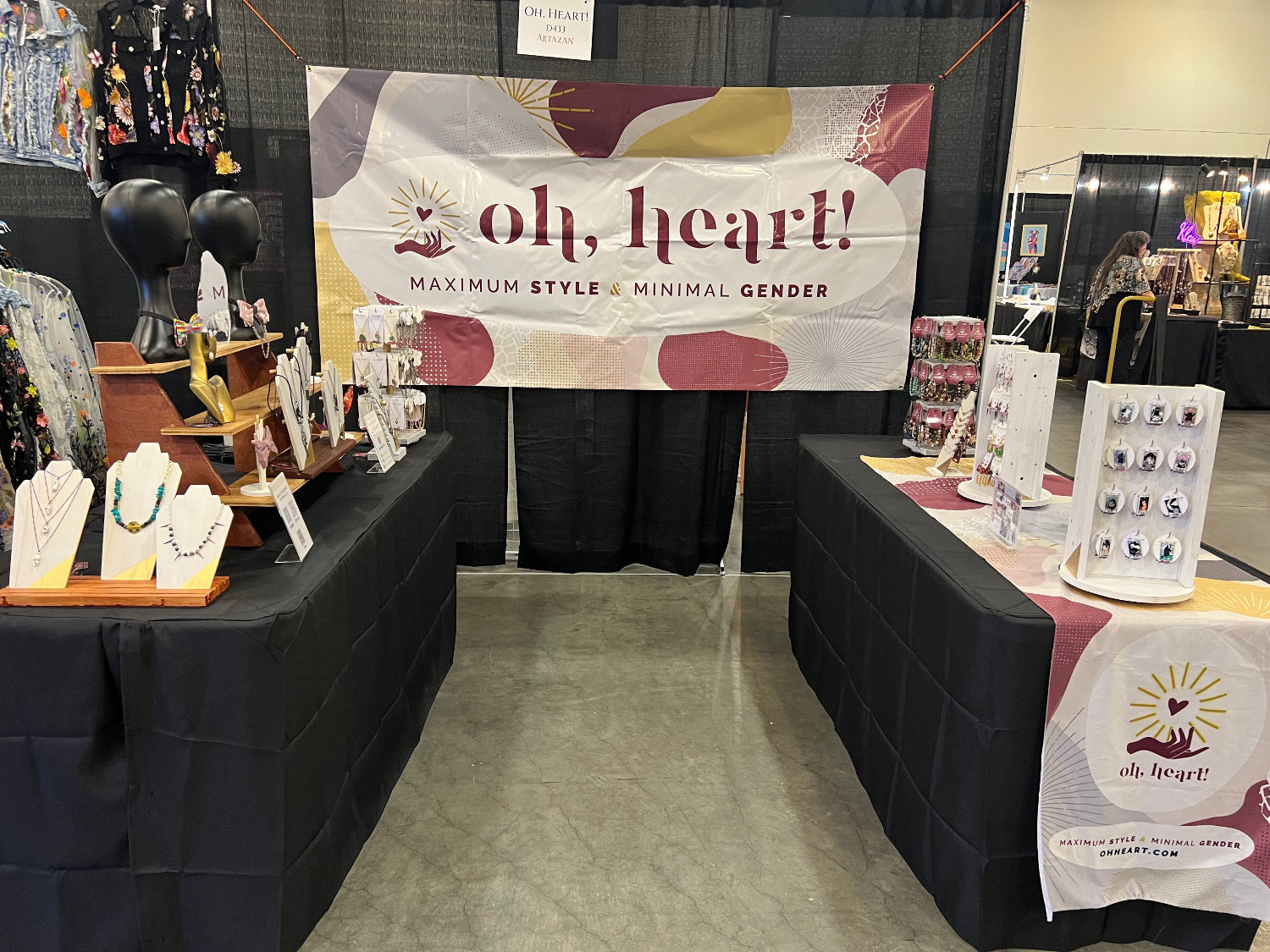 Photograph of Oh, Heart!'s vendor booth. Jewelry is displayed on multiple tables. A sign is hung up with the Oh, Heart! logo and the tagline "maximum style & minimal gender"