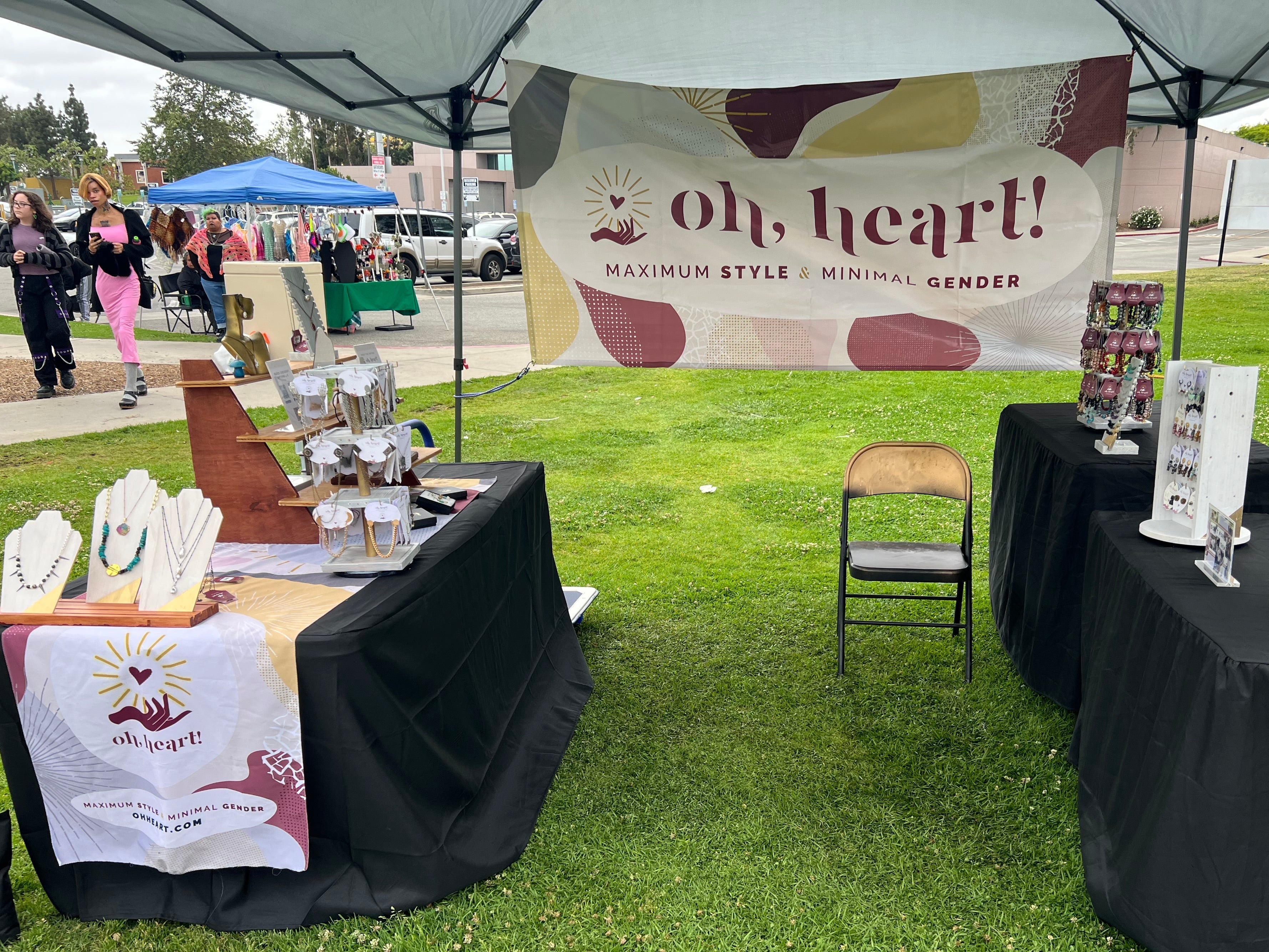Photograph of Oh, Heart!'s vendor booth at Queer Mercado in May, 2025. Jewelry is displayed on multiple tables. A sign is hung up with the Oh, Heart! logo and the tagline "maximum style & minimal gender"