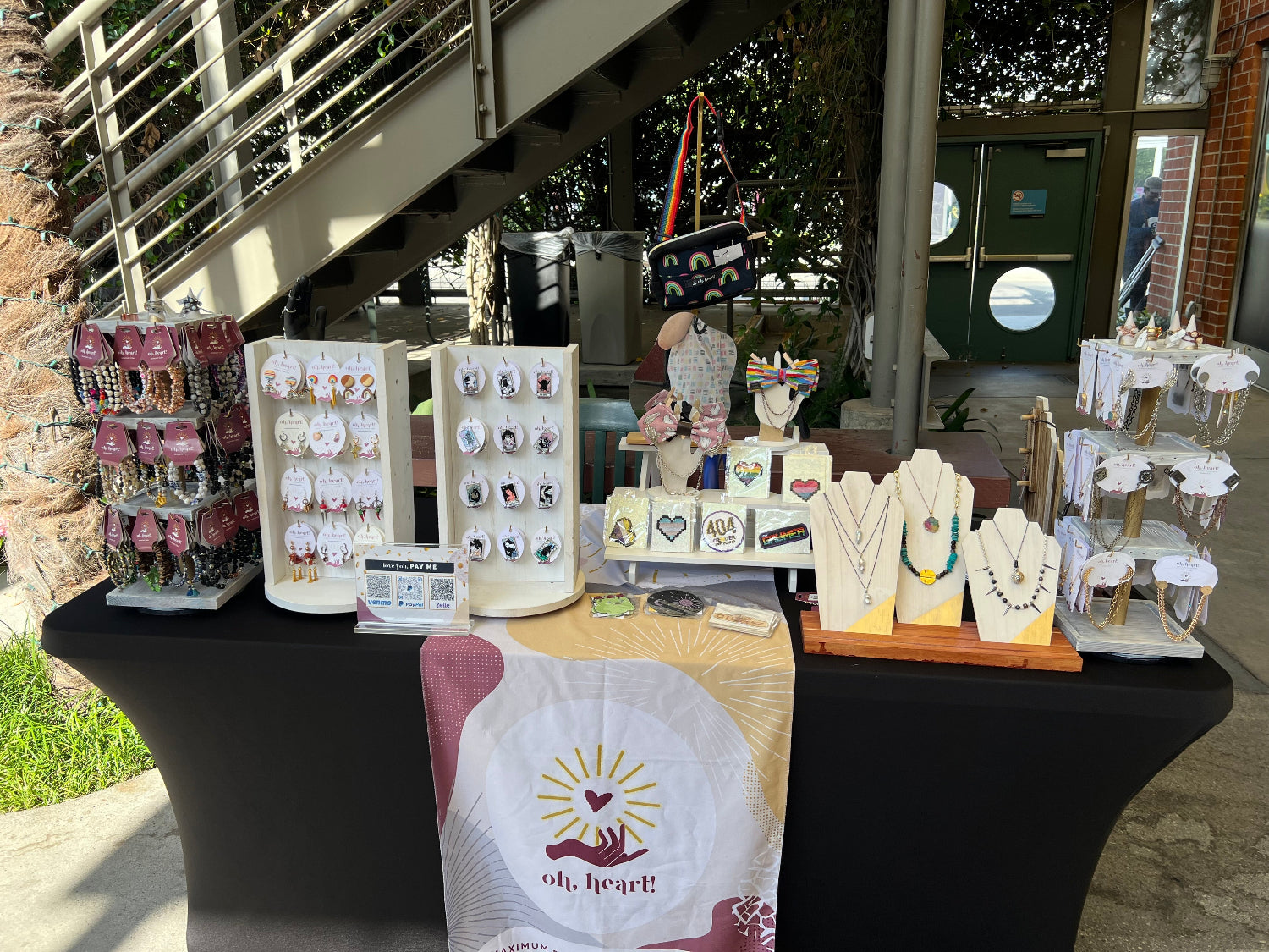 Photograph of Oh, Heart!'s vendor table at the WeHo Pride Art Festival in May 2025. Jewelry and fashion accessories are displayed on a table. A table runner is laid across the table with the Oh, Heart! logo.
